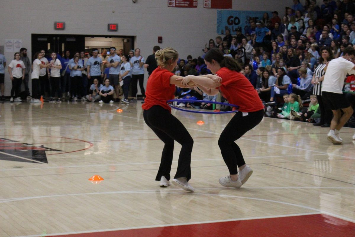  High school English teachers Emma Stiffler and Kelly Porter hold arms staying as close as possible to not let the hula hoop drop to the floor during this relay race. 
¨When Ms. Porter and I first found out that we were in the hula hoop challenge, we talked about how we might approach it during the practice before the Battle of the Buildings,” Stiffler said. “We first said that we were going to say the name of the person's direction we were going. When we got to the challenge, we saw a few others go before us. From what we saw, we decided to shift our plan a bit. We held hands, stayed low, and still used the communication tactic we originally came up with. All of these factors combined helped us zig-zag through the cones quickly and without dropping the hula hoop. It was a bit nerve racking as we were trying to get to the other side of the court as fast as we could, but having Ms. Porter, who is also a friend of mine, I think contributed to our success.¨

